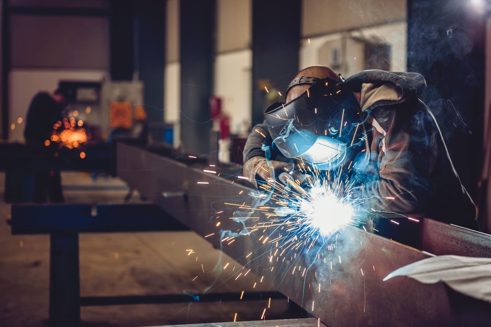 Welder in a welding helmet running a weld along a steel beam in a fabrication workshop, sparks flying