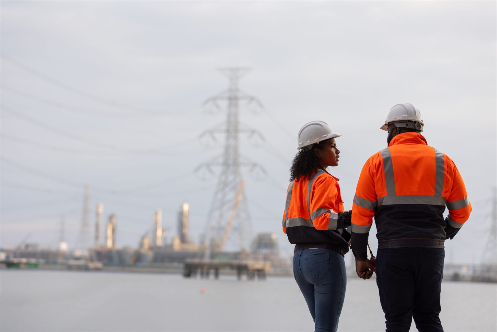 Two tradespeople in orange hi-vis jackets and white hard hats looking across an industrial waterfront towards transmission towers