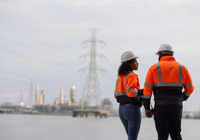 Two workers in orange safety vests and helmets stand together near a power plant with transmission towers in the background, symbolizing teamwork, industrial safety, and energy sector operations